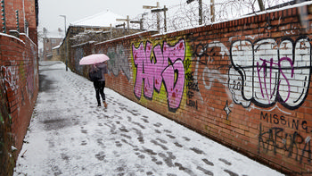 Alley snow walker This urban photograph captures the scene on Union Walk in Chesterfield, Derbyshire, United Kingdom, during a winter morning. The main subject is a person walking through the alleyway, holding a pink and white umbrella to shield themselves from the falling snow. Snow covers the path and the surrounding architecture, which consists of brick walls adorned with colorful graffiti. The snow and footprints highlight the wintry conditions of the morning. The image features elements of urban architecture typical of this part of Chesterfield, providing a snapshot of daily life in the United Kingdom during winter.